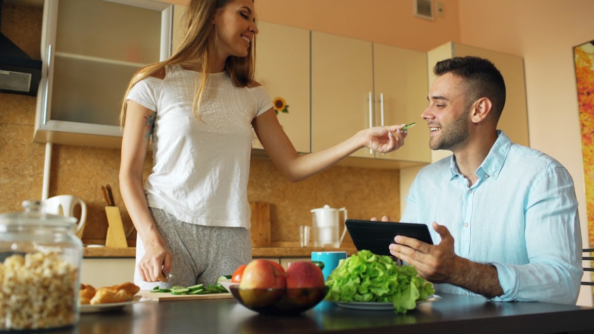 🎬 FULL 4K VIDEO for this still is in my bio link! ⬇️ Attractive couple chatting in the kitchen early morning. Beautiful woman feed her husband while cooking breakfast at home